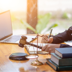 photo of a desk arrayed with items - three books stacked, scales of justice, a person's hand holding a gavel in the air, other hand holding a pen poised over a pad of paper, an open laptop in the background