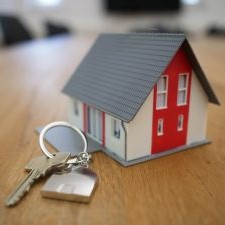 Image of a tiny house (grey sloped triangular roof, white walls, with a red vertical stripe accent at the front) sitting on a wooden table-top, with a single key on a keychain ring resting beside the house