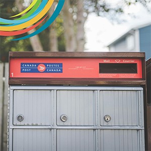 Partial image of a community mailbox, showing the mail slot on a red background, and closed, grey mailboxes. A blue house and large tree are blurred in the background. Photo by Stephen Andrews: https://www.pexels.com/photo/red-and-gray-mailbox-9900013/