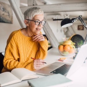 Women at desk looking at laptop with pen in her hand with notebook beside her 