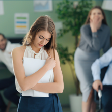 Sad woman standing in office while colleagues laugh at her