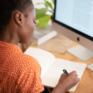 Professional woman working at desk writing in book with computer screen in front of her