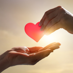 Picture of two hands with one hand passing a red paper heart to the other hand. The sun is shining in the background