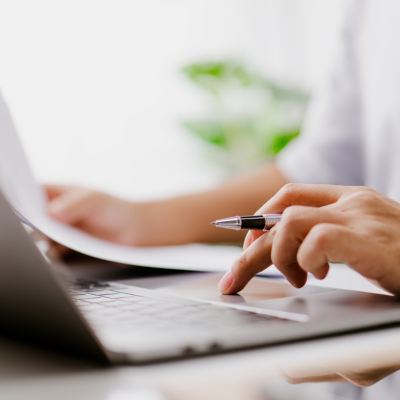 Picture of hand working on laptop with one hand holding a pen and one holding paper
