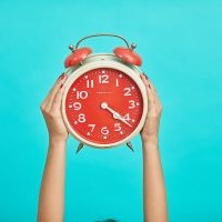 Hands holding up an alarm clock with a red face, white arms and white numbers in front of a light blue background;