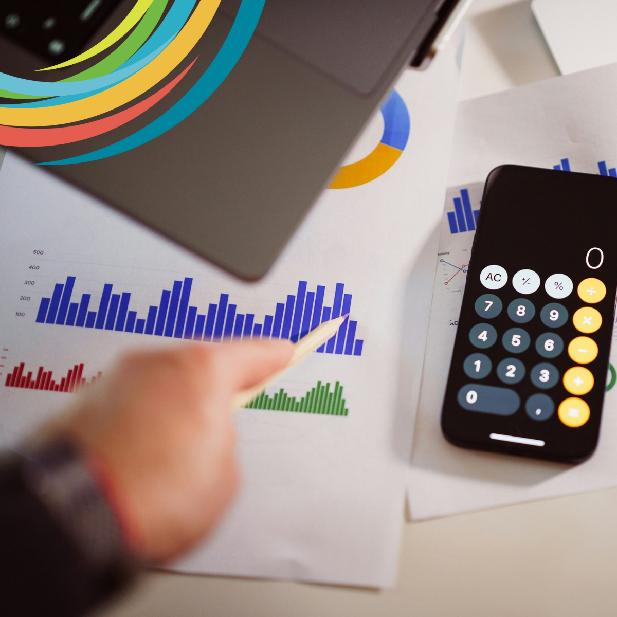 Table with several papers showing charts, graphs, and calendars. On top of the papers is a phone calculator, and a person's blurry hand is holding a pencil which points at the calculator. Photo by Jakub Żerdzicki on Unsplash