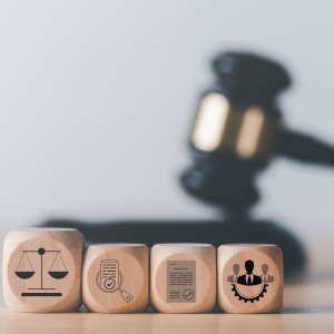  In the foreground are four wooden blocks with simple icons on each. A set of scales, a magnifying glass over a document, a letter with a seal affixed, and a group of three men. In the background is a blurred judge's gavel resting on the gavel block.