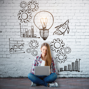 Young woman sitting on ground with laptop with images of lightbulb, wheels, graphs drawn on wall behind her